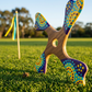 Boomerang quadripale Balabalaa aux pointillés aborigènes violet, jaune et turquoise, posé sur pelouse avec drapeau en fond