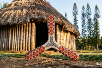Three-blade Kanak boomerang with red and orange diamond tribal patterns, posed in front of a traditional New Caledonian hut