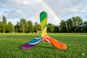 Colorful three-blade boomerang with rainbow swirl pattern, posed on green grass with a rainbow in the sky