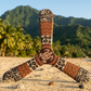 Boomerang tripale Tahiti aux motifs polynésiens brun et beige avec tortue et hibiscus, posé sur une plage de sable blanc