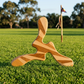 Boomerang tripale Yallingup M en bois naturel aux veines dorées, posé sur pelouse australienne avec drapeau en arrière-plan