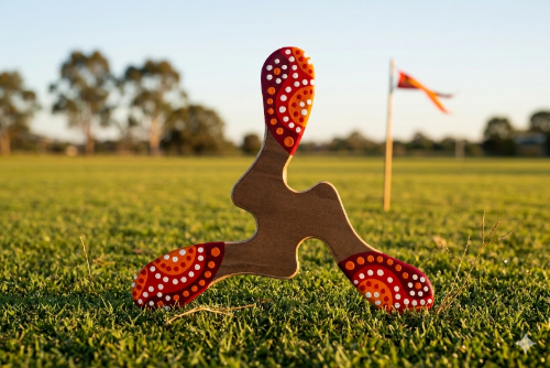 Boomerang tripale Yallingup bois naturel et extrémités rouge à pointillés aborigènes, posé sur pelouse au coucher de soleil