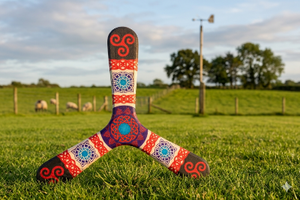 Boomerang tripale Celtic aux motifs triskel et entrelacs celtiques rouge, violet et turquoise, posé dans un pré irlandais verdoyant